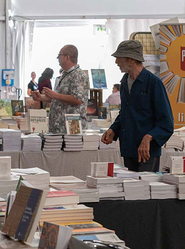 Des piles de livres sont posées sur des tables, deux hommes sont tournés vers la gauche